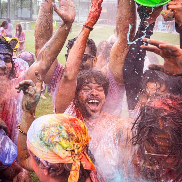 A group of students laugh and splash water on each other while covered in orange, yellow, purple and blue powder at the on-campus Holi celebration.